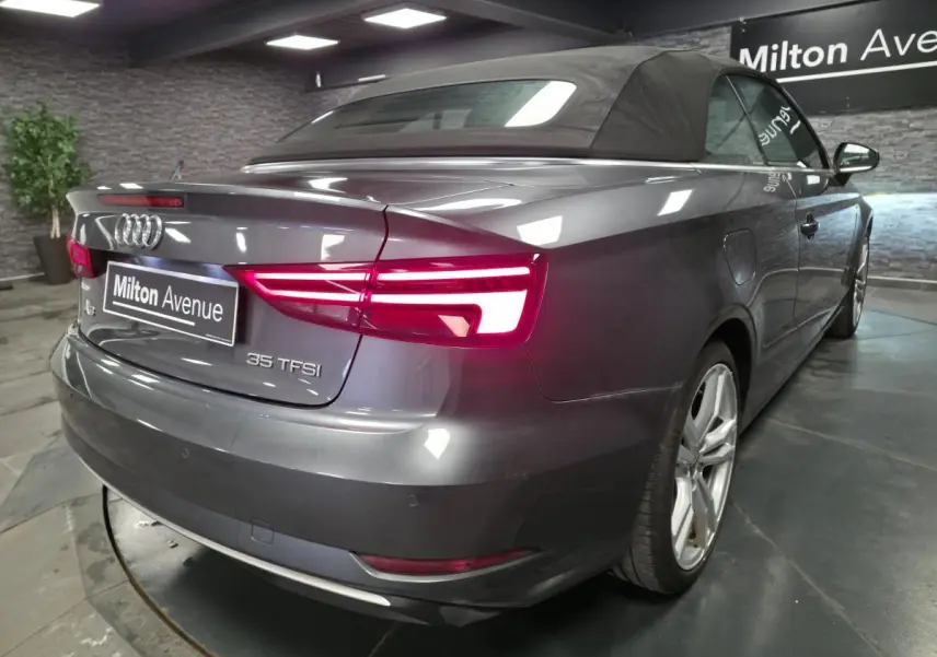 Audi A3 Cabriolet gris vue 3/4 arrière droit avec capote noire et feux arrière allumés dans un showroom.