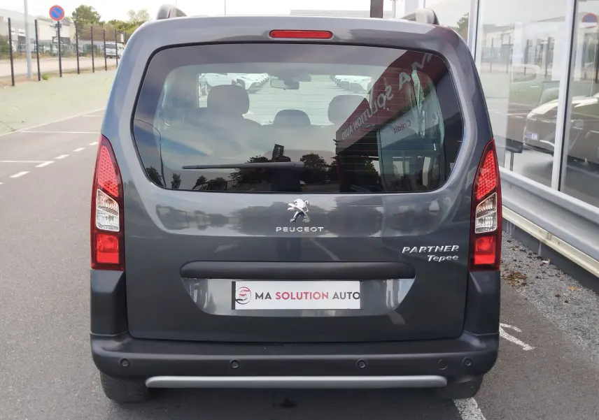Vue arrière d'un Peugeot Partner Tepee gris foncé, avec logo et badges visibles, stationné devant un bâtiment vitré.