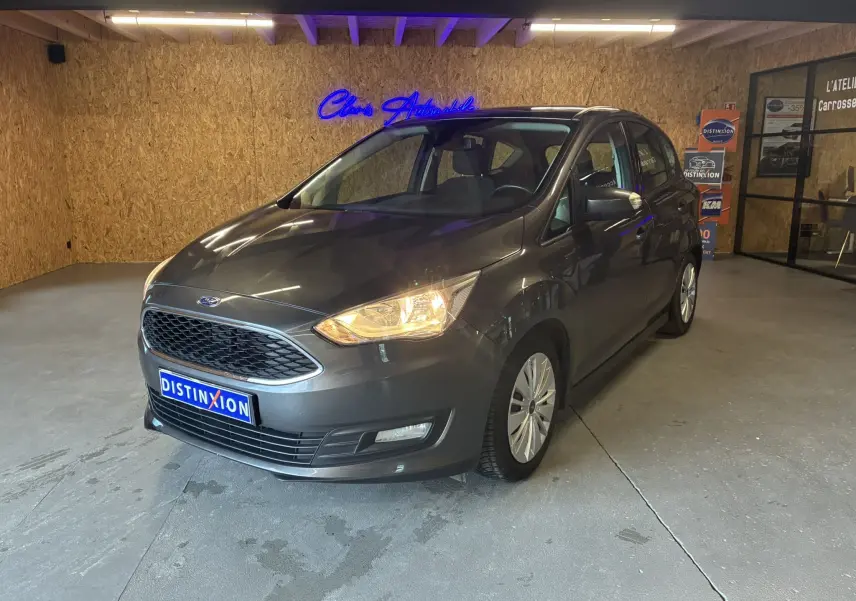 Ford C-MAX gris 3/4 avant droit en intérieur avec phares allumés et calandre hexagonale distinctive.