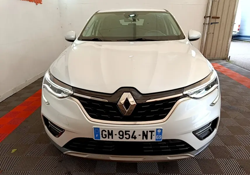 Vue frontale d'une Renault Arkana blanche 2023 avec calandre noire et phares LED allumés dans un garage.