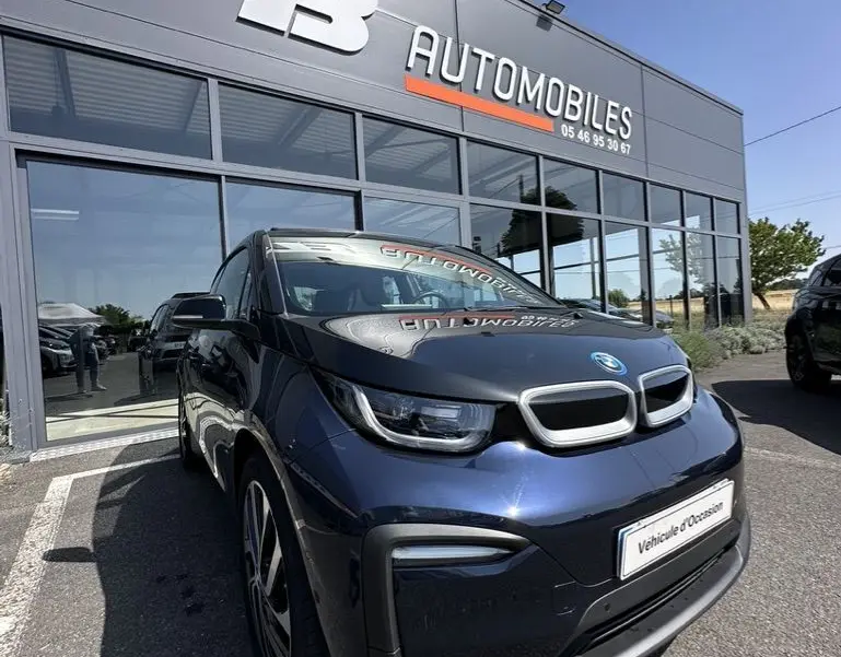 BMW i3 bleue vue en 3/4 avant droit, garée devant un concessionnaire FB Automobiles par temps clair.