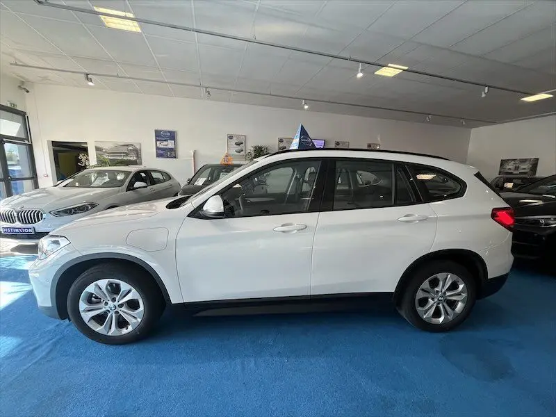 BMW X1 XDRIVE 25E blanc alpin en vue latérale gauche, hybride rechargeable exposée en showroom lumineux.