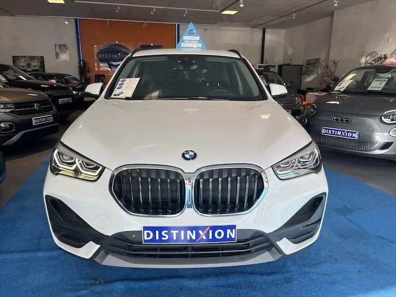 Vue frontale d'un BMW X1 XDRIVE blanc alpin en showroom avec calandre noire et phares LED allumés.