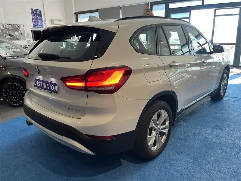 BMW X1 XDRIVE blanc alpin vue 3/4 arrière droit en intérieur avec feux arrière allumés et plaque Distinxion.