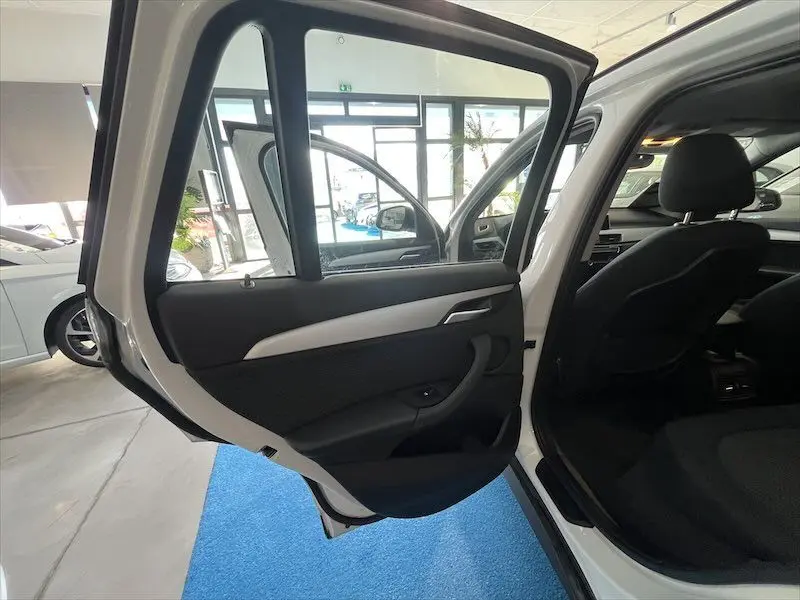Vue intérieure côté arrière gauche d'une BMW X1 XDRIVE blanche avec porte ouverte montrant les sièges noirs et garnitures de porte.