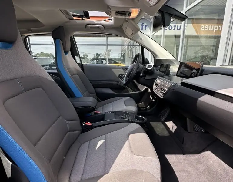 Vue intérieure côté conducteur de la BMW i3 noire 2022, sièges gris avec surpiqûres bleues et toit ouvrant panoramique.