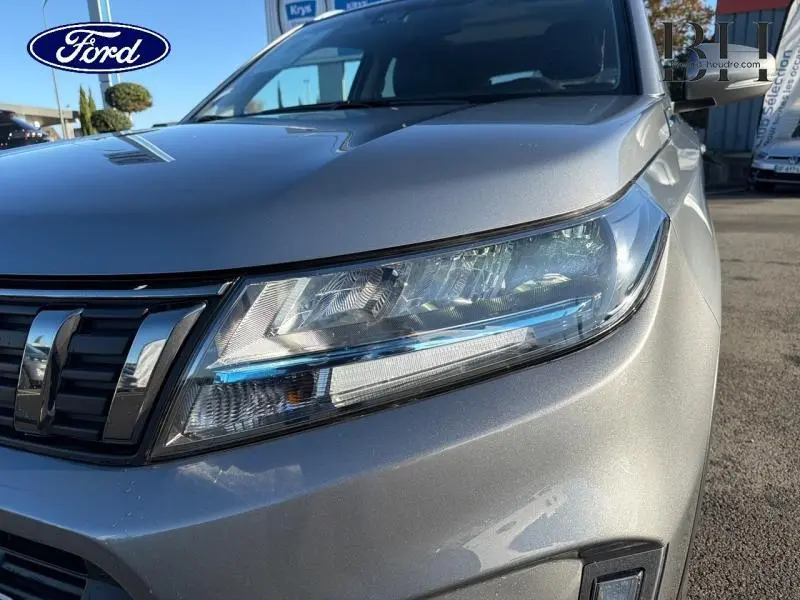 Gros plan sur le phare avant droit du Suzuki Vitara Galactic Gray métallisé avec calandre chromée sous lumière naturelle.