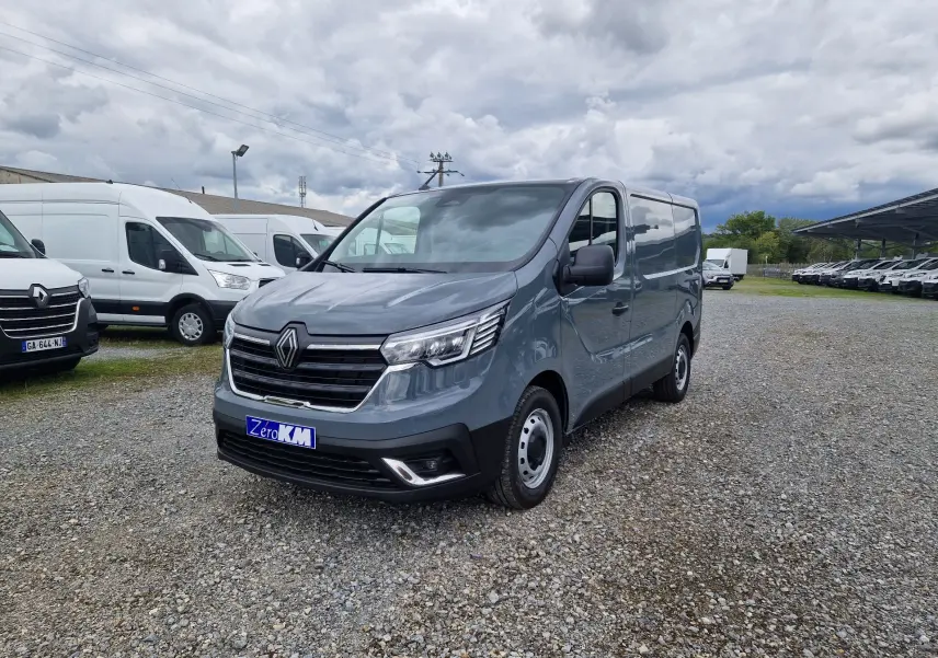 Renault Trafic Fourgon gris urbain vu en 3/4 avant droit sur un parking avec plusieurs utilitaires blancs en arrière-plan.