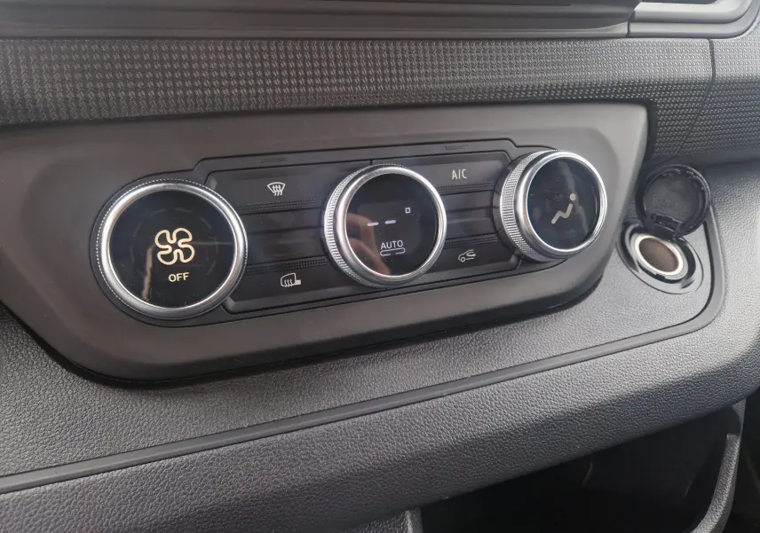 Gros plan sur la console de climatisation du Renault Trafic Fourgon gris urbain, montrant les trois boutons rotatifs et une prise 12V ouverte.