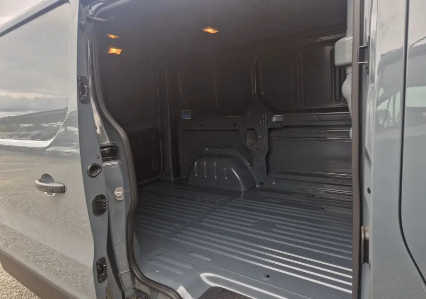 Vue latérale gauche sur l'ouverture du fourgon Renault Trafic L1H1 gris urbain, montrant l'espace de chargement vide et éclairé.