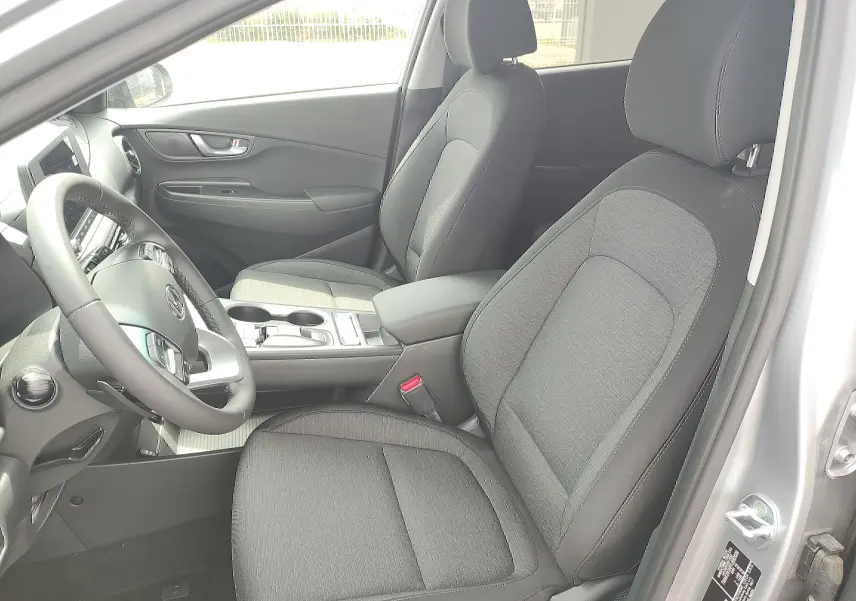 Intérieur avant du Hyundai Kona Electric 2025, sièges tissu noir et console centrale visible côté conducteur.