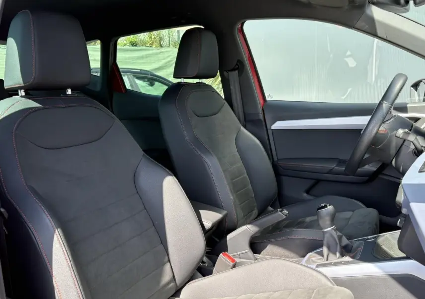 Intérieur avant du SEAT Arona TSI 110 FR 2024, sièges en suédine noire avec surpiqûres rouges, volant et levier de vitesse visibles.