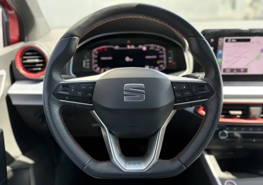 Volant sport noir avec surpiqûres rouges du SEAT Arona TSI 110 FR 2024, tableau de bord digital et écran tactile en arrière-plan.