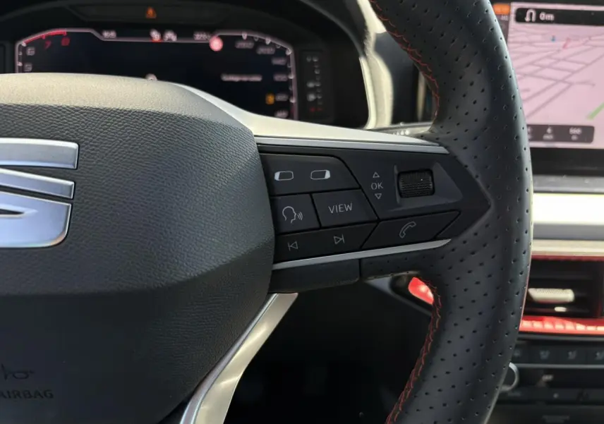 Gros plan sur le volant perforé noir avec surpiqûres rouges du SEAT Arona TSI 110 FR, tableau de bord numérique en arrière-plan.