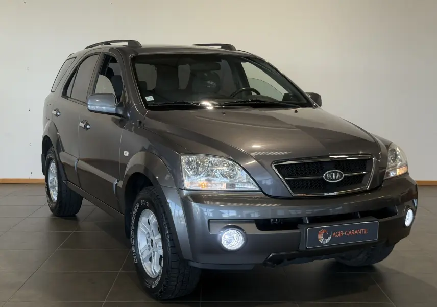 SUV KIA Sorento gris 2005 vu en 3/4 avant droit avec phares et antibrouillards allumés en intérieur showroom.