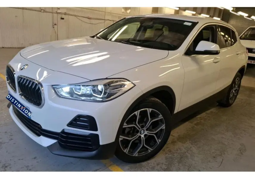 BMW X2 blanc vue 3/4 avant droit, jantes alliage 18'', rétroviseurs électrochromes et phares allumés.
