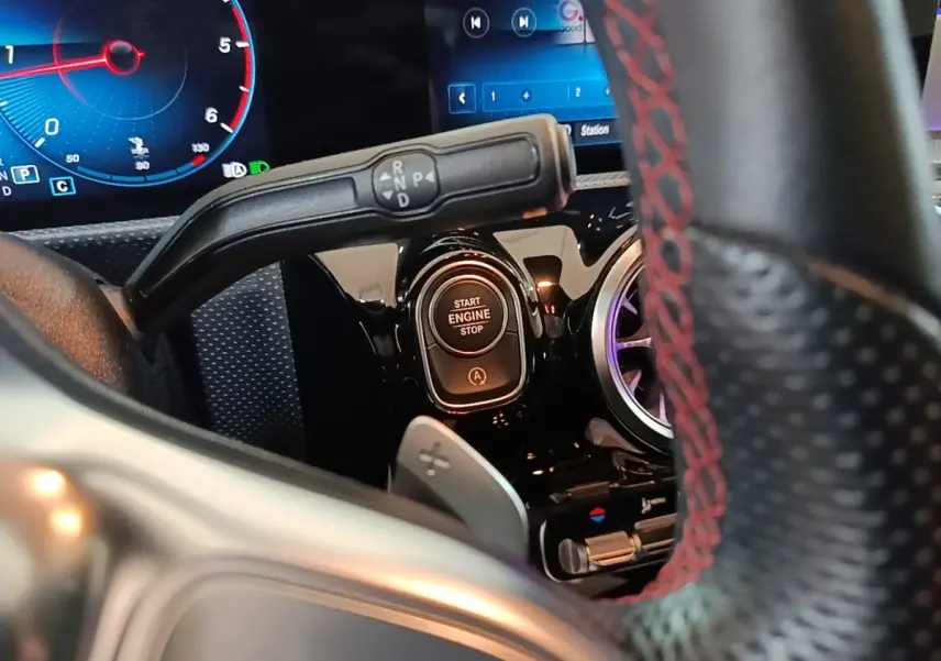 Gros plan intérieur sur le bouton démarrage et levier de boîte automatique d’une Mercedes CLA 200 d AMG Line noire, volant cuir noir à surpiqûres rouges visible.