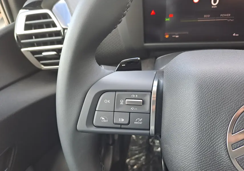 Gros plan sur les commandes à gauche du volant d'une Citroën C4 hybride 2025, avec tableau de bord numérique en arrière-plan.