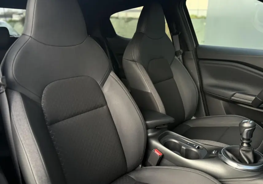Vue intérieure côté avant droit du Nissan Juke 2025 avec sièges noirs en cuir et tissu, console centrale et levier de vitesses manuel.