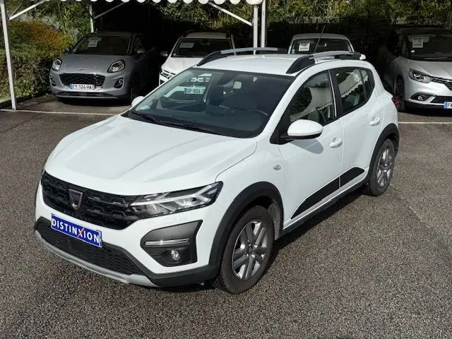 Vue 3/4 avant droite d'une Dacia Sandero Stepway blanche avec barres de toit et protections noires sur les passages de roues.