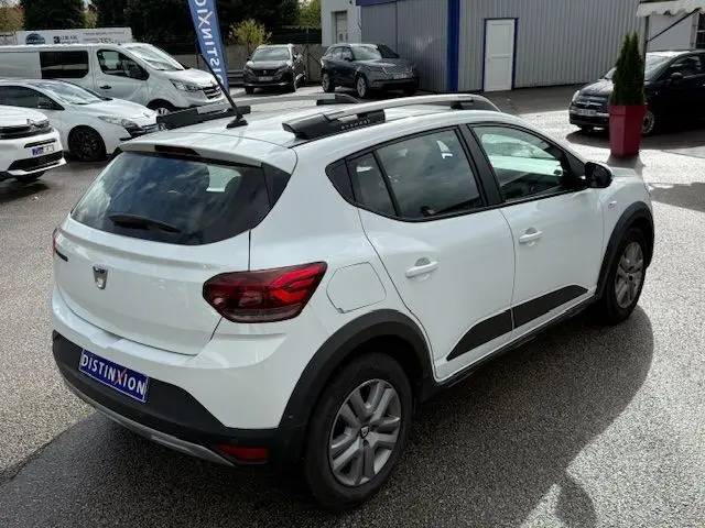 Vue 3/4 arrière droite d'une Dacia Sandero Stepway blanche avec barres de toit et protections noires sur les bas de caisse.