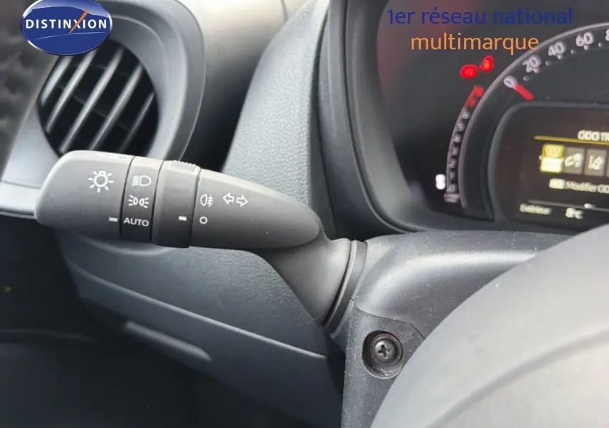 Gros plan sur la commande des phares et clignotants à gauche du volant d’une Toyota Aygo X noire, tableau de bord partiellement visible.