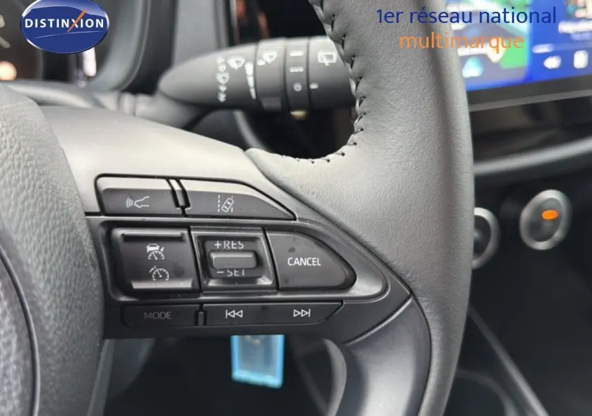 Gros plan sur les commandes au volant en cuir noir de la Toyota Aygo X 2025, avec tableau de bord flou en arrière-plan.