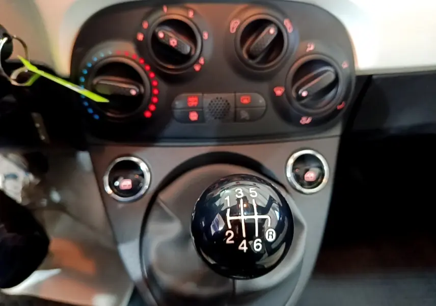 Gros plan sur le levier de vitesses manuel noir et la console centrale avec commandes de climatisation d'une Fiat 500 blanche.