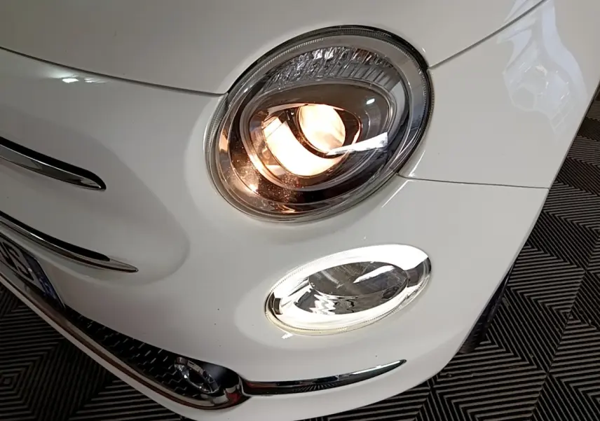Gros plan sur le phare avant droit allumé d'une FIAT 500 blanche, mettant en valeur les détails chromés du pare-chocs.