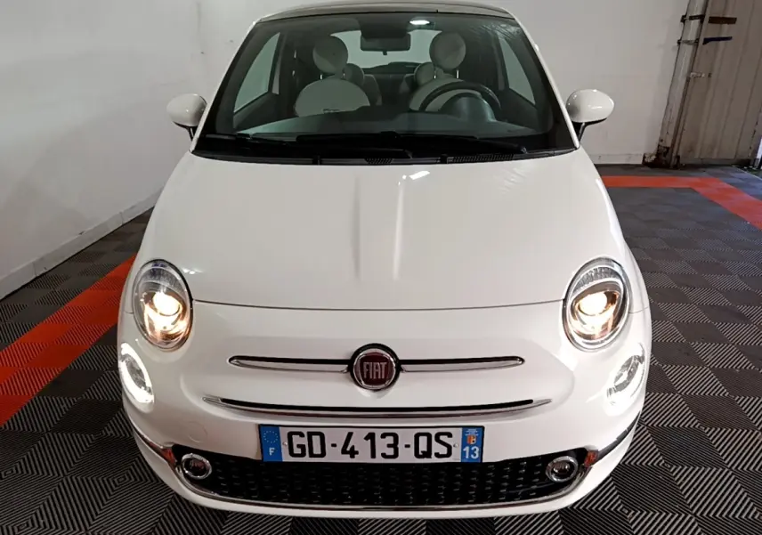 Vue avant d'une FIAT 500 blanche avec phares allumés, logo centré et plaque d'immatriculation française visible.