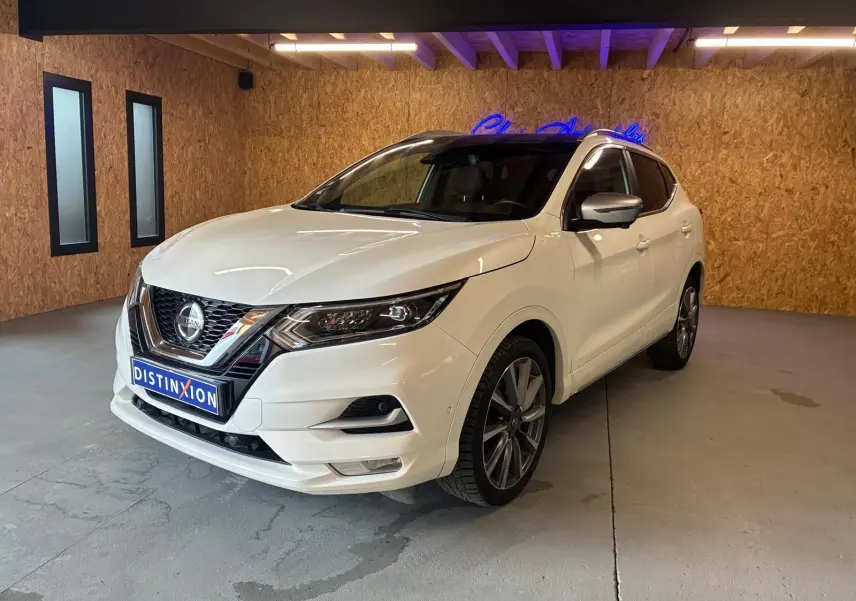 Nissan Qashqai blanc en 3/4 avant droit, intérieur visible noir, dans un showroom avec mur en bois et néon bleu.