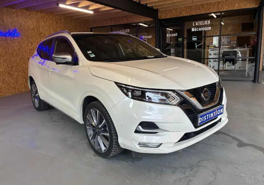 Nissan Qashqai blanc en 3/4 avant droit dans un garage, avec jantes alliage et toit panoramique noir.