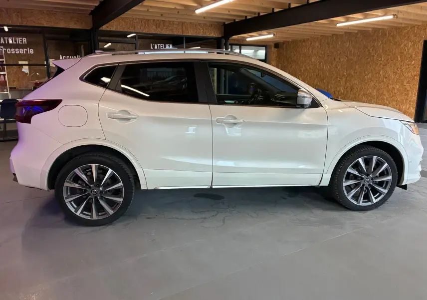 Vue de profil côté gauche d'un Nissan Qashqai blanc 2021 dans un garage, avec jantes alliage et vitres teintées.