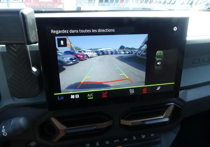 Écran tactile intérieur du Dacia Duster blanc 2025 montrant la caméra de recul avec vue arrière sur un parking.