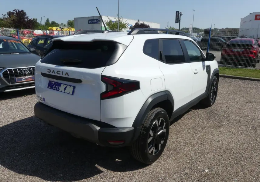 Vue 3/4 arrière droite d'un Dacia Duster blanc 2025 avec jantes alliage noires et barres de toit noires.