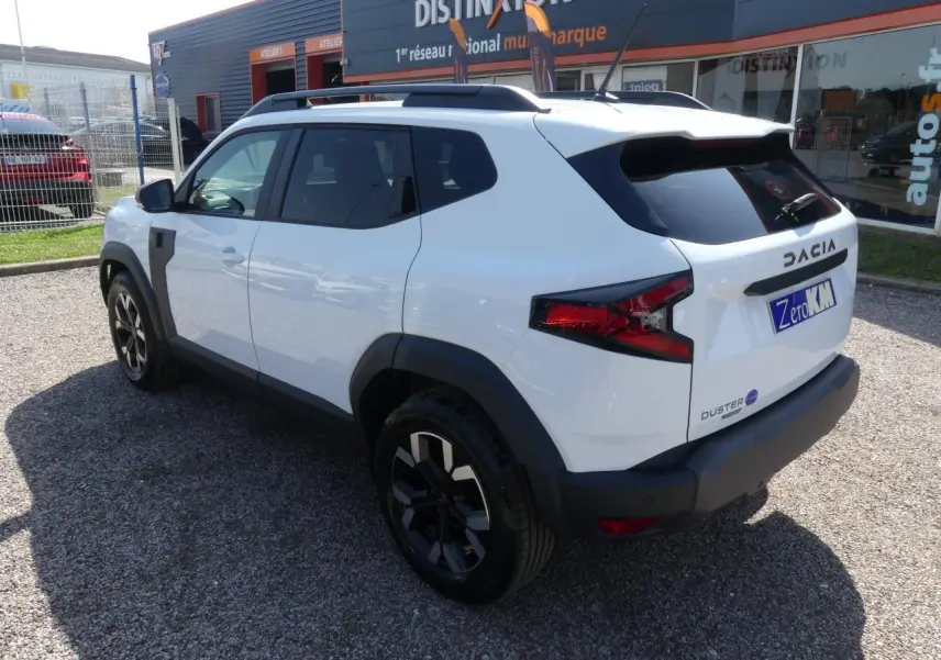Vue 3/4 arrière droite d'un Dacia Duster blanc 2025 avec jantes alliage noires et barres de toit noires.