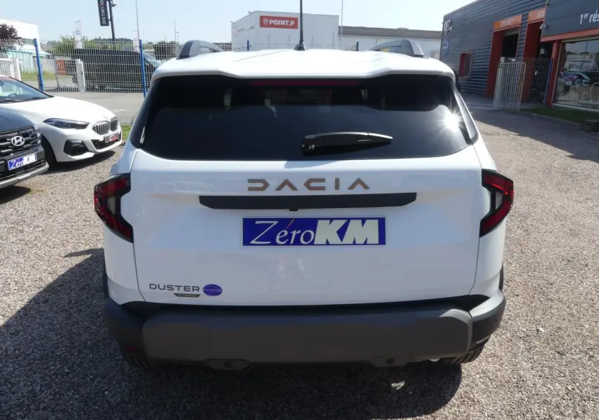 Vue arrière d'un Dacia Duster blanc avec logo brun cuivré et plaque d'immatriculation Zero KM visible.
