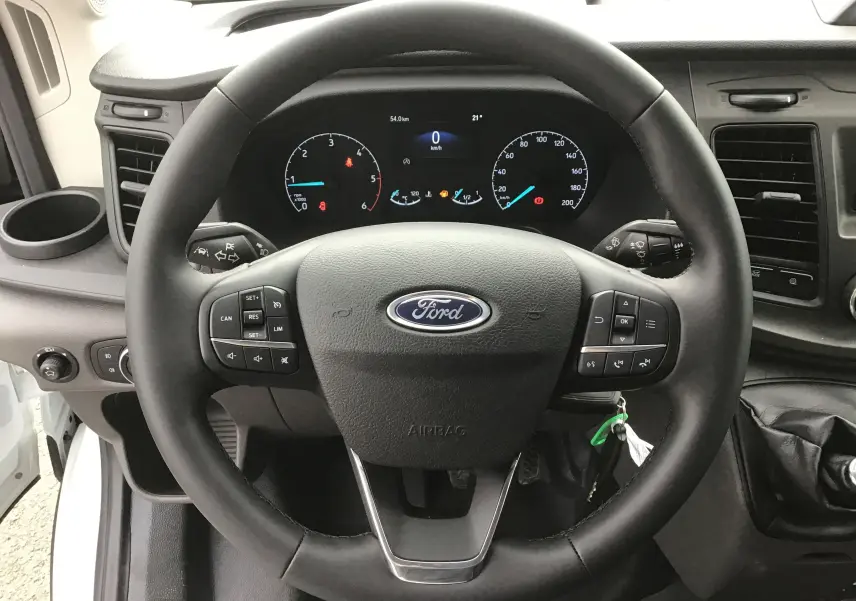 Vue rapprochée du volant noir Ford dans l'habitacle du Transit Châssis Cabine blanc, avec tableau de bord numérique allumé.