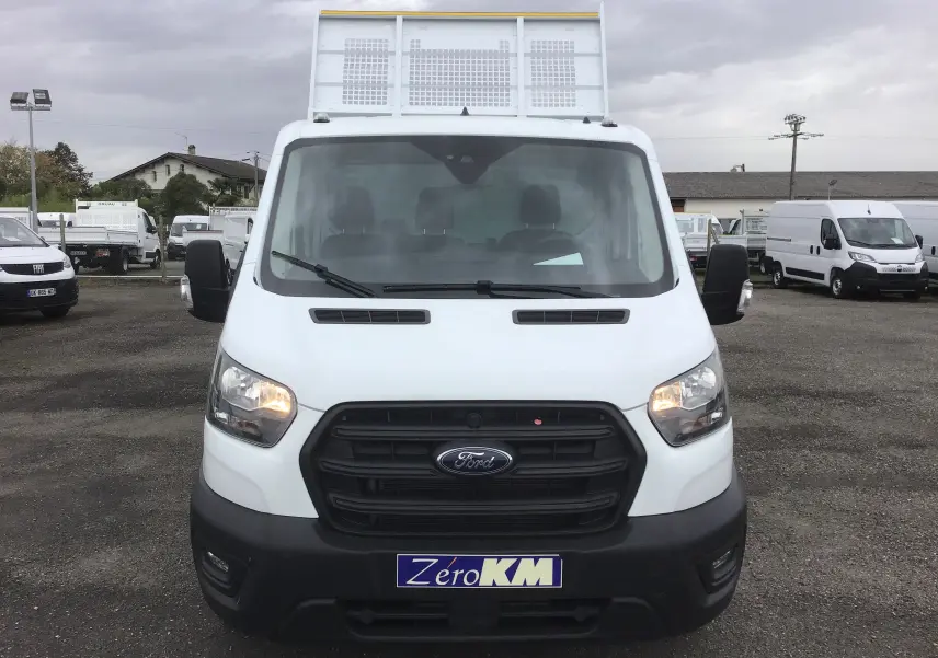 Vue frontale d'un Ford Transit blanc avec benne et coffre, phares allumés sur un parking extérieur.