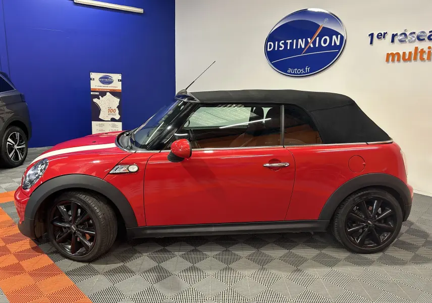 Profil droit d'une MINI Cabriolet Cooper S rouge avec capote noire et jantes noires en intérieur showroom.