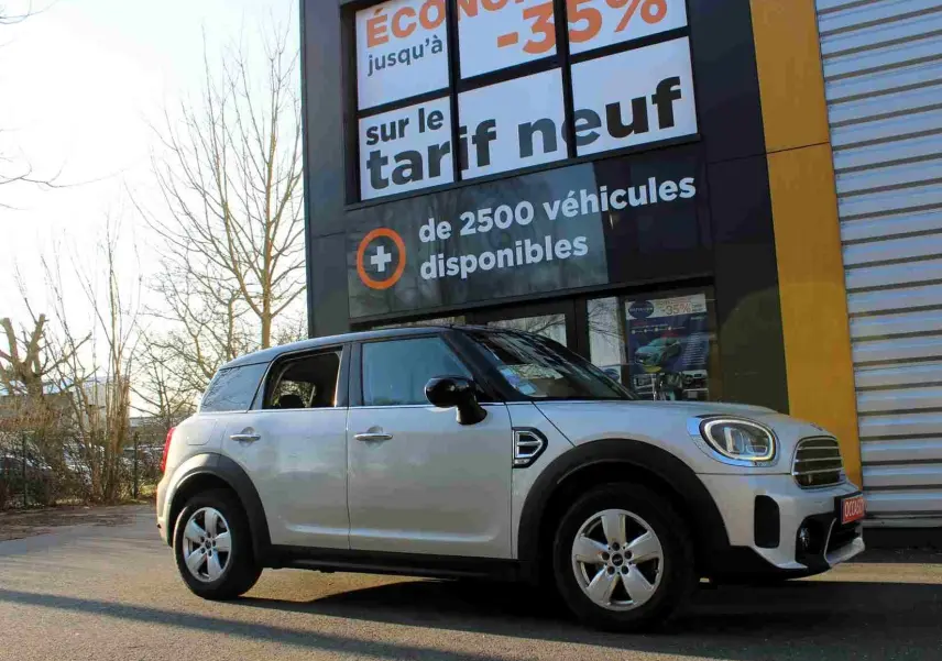 Vue latérale gauche d'un MINI Countryman gris perle métal avec toit noir, stationné devant une enseigne commerciale.