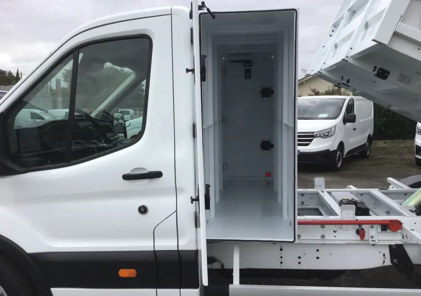 Vue latérale gauche d'un Ford Transit blanc avec benne relevée et coffre de rangement ouvert sur châssis cabine.