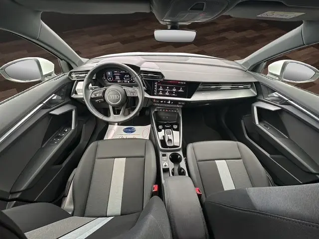 Vue intérieure avant de l'Audi A3 Sportback 2024 avec tableau de bord digital, volant cuir multifonctions et sièges tissu noir.