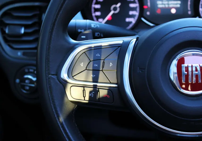 Gros plan sur le volant cuir noir de la Fiat 500X 2019, avec commandes multifonctions et logo FIAT rouge au centre.