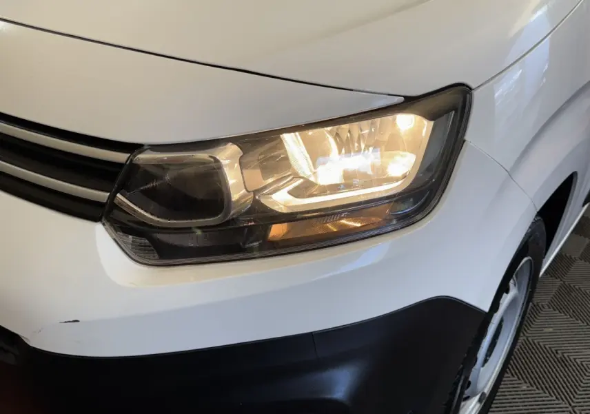 Gros plan sur le phare avant droit allumé d’un Citroën Berlingo blanc avec pare-chocs noir.