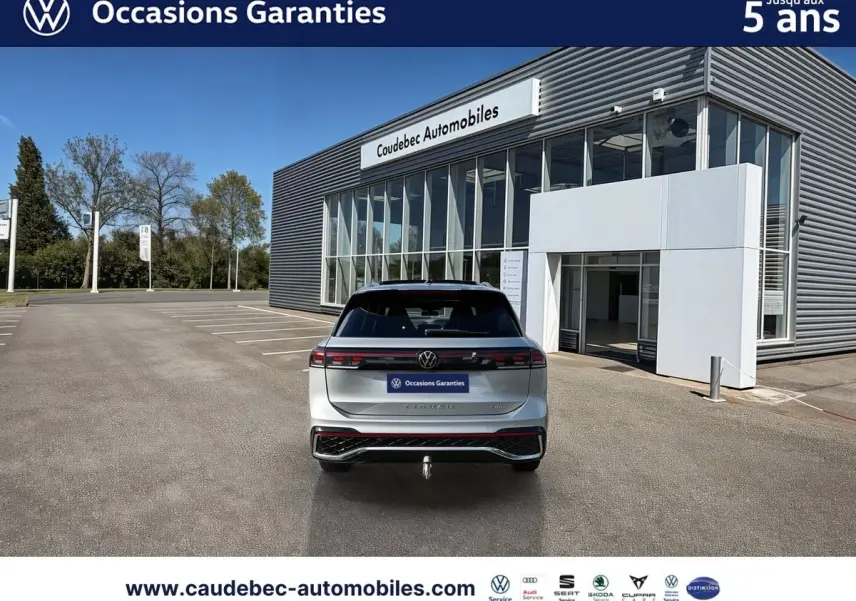 Vue arrière d’un Volkswagen Tiguan eHybrid 2025 argent Dolomite avec hayon électrique et crochet d’attelage escamotable, devant un bâtiment.
