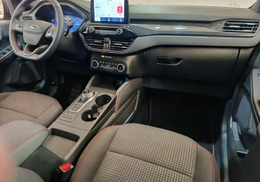 Intérieur du Ford Kuga 2024 St Line, vue côté conducteur, tableau de bord noir avec écran tactile et sièges tissu gris à surpiqûres rouges.