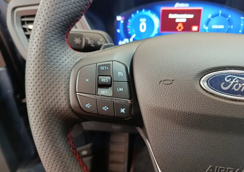 Gros plan sur le volant perforé avec surpiqûres rouges du Ford Kuga 2024, tableau de bord numérique bleu en arrière-plan.