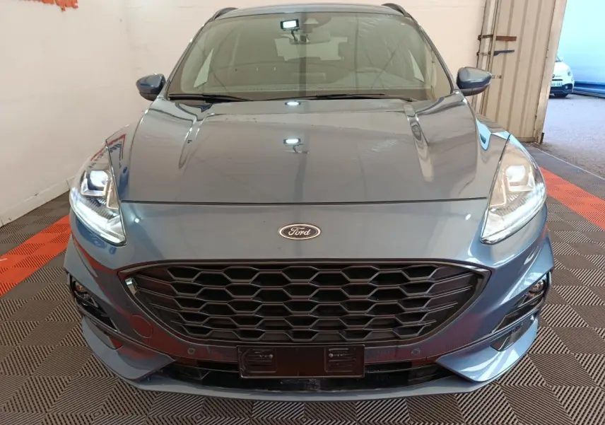 Vue frontale rapprochée du Ford Kuga bleu 2024 avec calandre noire et phares allumés en intérieur.