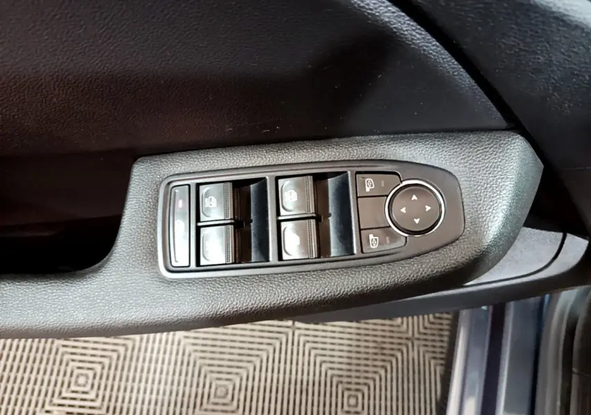 Gros plan sur les commandes des vitres électriques et rétroviseurs sur la porte côté conducteur d’une Renault Clio gris foncé.